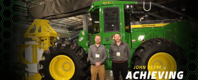 Lee Gulbrandson and Tyle Chrstensen pose in front of John Deere peice of logging equipment while at an Acheiving Excellence reception.